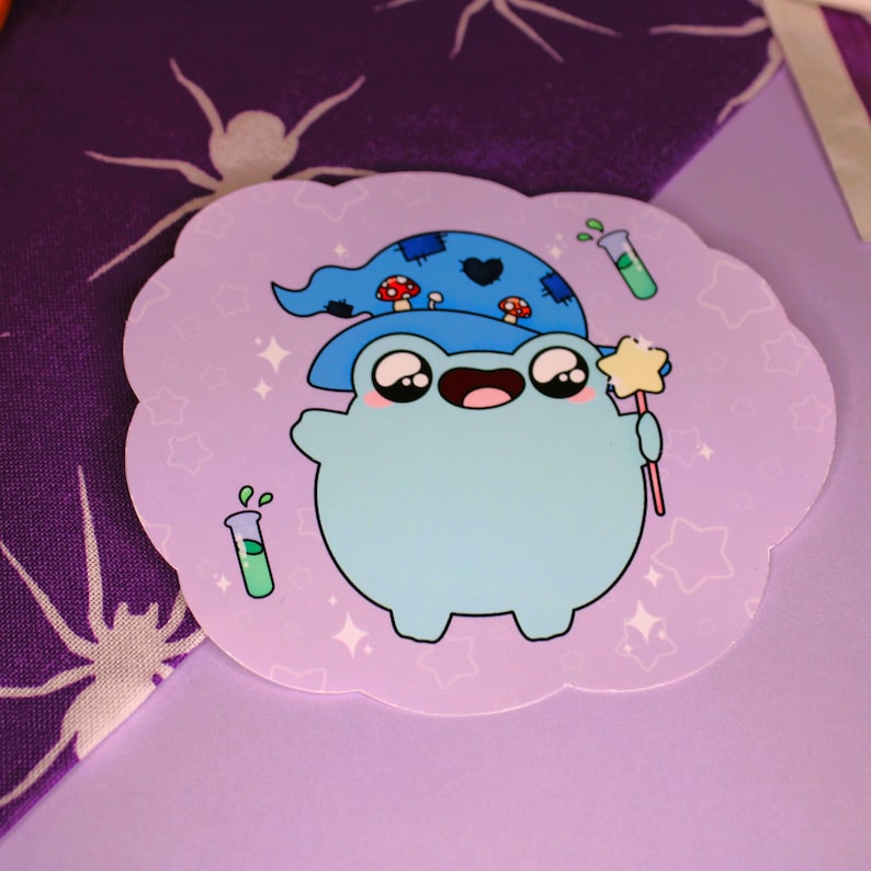Peut inclure: Une grenouille bleue portant un chapeau de sorcier bleu et noir avec des champignons sur le dessus, tenant une baguette avec une &eacute;toile au sommet. La grenouille est entour&eacute;e d'&eacute;toiles et de tubes &agrave; essai sur un fond violet clair.
