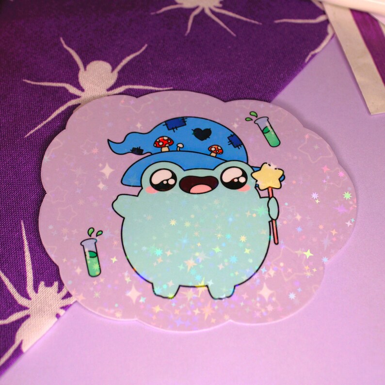 Peut inclure: Un autocollant holographique repr&eacute;sentant une grenouille de dessin anim&eacute; portant un chapeau de sorcier bleu et noir avec des champignons sur le dessus. La grenouille tient une baguette avec une &eacute;toile au sommet et est entour&eacute;e d'&eacute;toiles et de paillettes.