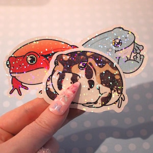 May include: Three glittery frog stickers in red, blue, and brown with black outlines. The red frog has a bright red body and the blue frog has a light blue body. The brown frog has a brown and tan pattern. The stickers are on a light blue background with white polka dots.