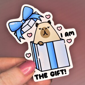 Op de afbeelding: Een sticker met een cartoonillustratie van een hond in een blauwe en witte cadeaudoos. De hond kijkt uit de doos en de tekst "Ik ben het cadeau!" staat onder de doos.