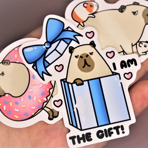 Op de afbeelding: Een set van vier stickers met een mopshond in verschillende poses. E&eacute;n sticker toont de mopshond met een roze donut, een andere toont de mopshond in een blauwe cadeaudoos met een blauwe strik, en de andere twee stickers tonen de mopshond met de tekst "Ik ben het cadeau!" en een hartje.