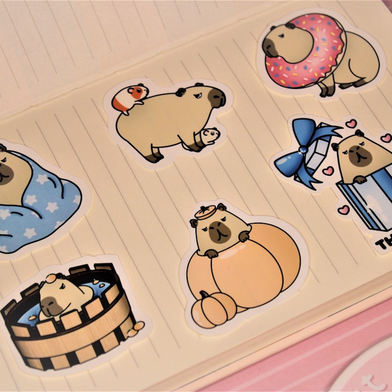 Op de afbeelding: Een set van acht stickers met schattige cartoon capybara's in verschillende poses en settings. De stickers zijn gedrukt op wit papier en hebben een glanzende afwerking. De capybara's worden afgebeeld in verschillende activiteiten, waaronder het dragen van een donut, zitten in een pompoen en baden.