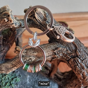 May include: A silver-toned keychain with a decorative knot and a heart-shaped clasp. The keychain has a ring with copper and green beads. The word "mine" is written in quotation marks at the bottom.