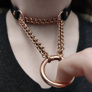 Op de afbeelding: Een roségouden kettingchoker met een grote roségouden ring eraan bevestigd. De choker is gemaakt van een gevlochten ketting en heeft een zwarte leren achterkant.