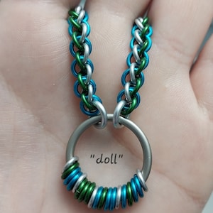 May include: A silver, green and blue chain necklace with a silver ring pendant. The ring is wrapped with silver, green and blue wire. The text "doll" is written on the pendant.