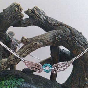 May include: A silver chain necklace with a delicate, openwork design of wings on either side of a teal-colored ring. The necklace is displayed on a piece of driftwood.