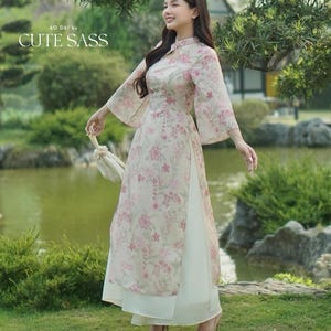 May include: A woman wearing a floral print Ao Dai dress with a high collar and flared sleeves. The dress is light pink with a floral pattern and the text "CUTE SASS". The dress is paired with nude heels.