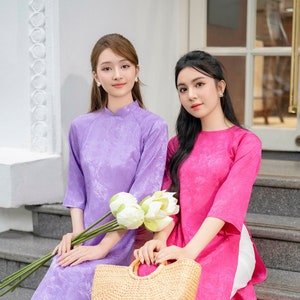 May include: Two women wearing traditional Vietnamese dresses, known as Ao Dai, in purple and pink. The women are sitting on a set of stairs with a woven straw bag. The woman in the purple dress is holding a bouquet of white lotus flowers.