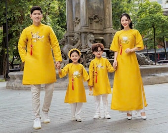 May include: A family of four wearing matching yellow traditional Vietnamese clothing with embroidered details. The family is standing in front of a stone fountain and a stone structure with columns. The text "AO DAI by CUTE SASS" is visible at the bottom of the image.