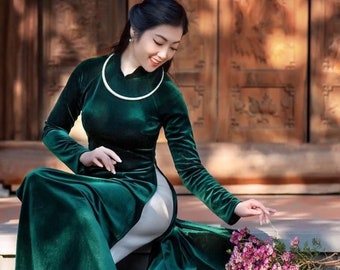 May include: A woman in a long, emerald green velvet dress with a white trim sits on a set of stone steps. She is holding a bouquet of pink flowers.