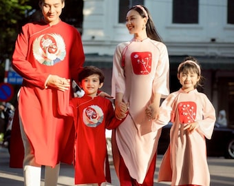 May include: A family of four wearing traditional Vietnamese clothing, known as Ao Dai, in red and pink colors. The father and son are wearing red Ao Dai with embroidered phoenix designs, while the mother and daughter are wearing pink Ao Dai with embroidered floral designs. They are walking on a street.