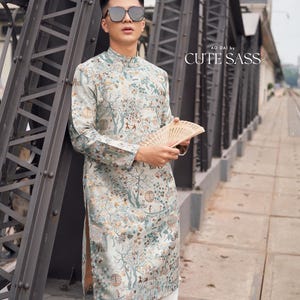 May include: A light green Ao Dai with a tree and figure pattern, paired with white pants and black boots. The outfit includes a hand fan. The text "AO DAI by CUTE SASS" is visible.