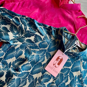 May include: Two silk blouses, one pink with a floral pattern and one teal with a leaf pattern. The teal blouse has a tag that reads "So darn cute, CUTE SASS".