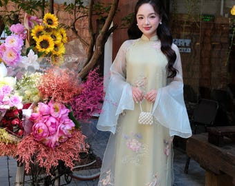 Pode incluir: Um Ao Dai vietnamita tradicional em verde claro com uma sobreposição transparente, mangas largas e delicados bordados florais. A peça é combinada com calças largas a condizer e complementada por uma pequena mala de mão com missangas brancas.