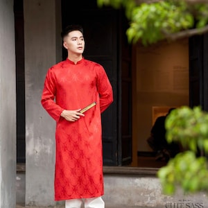 May include: A man wearing a red long-sleeved shirt with a traditional Vietnamese design, white pants, and black boots. He is holding a gold fan in his right hand. The shirt has a mandarin collar and is made of a silky fabric. The man is standing in front of a doorway with a wooden frame.