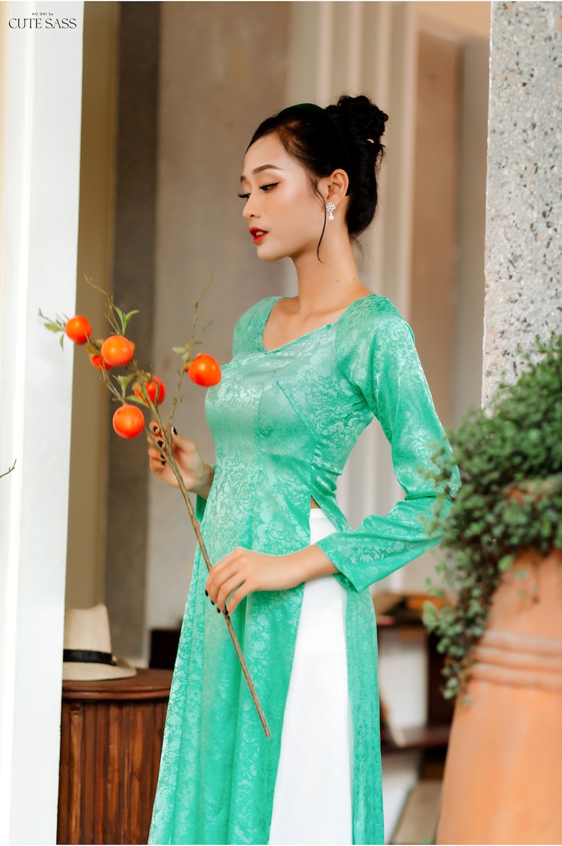 Op de afbeelding: Een vrouw draagt een tealgroene lange mouwen jurk met een witte rok en een bloemenpatroon. Ze houdt een tak vast met oranje fruit.