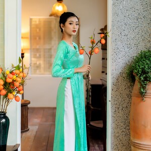 Op de afbeelding: Een vrouw in een teal zijden jurk met witte broek staat in een deuropening en houdt een tak met oranje fruit vast.