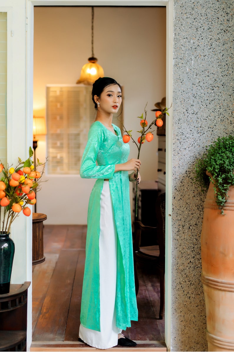 Op de afbeelding: Een vrouw in een teal zijden jurk met witte broek staat in een deuropening en houdt een tak met oranje fruit vast.