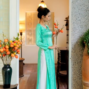 Op de afbeelding: Een vrouw in een turquoise zijden jurk met een witte rok en een tak met oranje fruit vasthoudt. De jurk heeft een traditionele Vietnamese stijl.