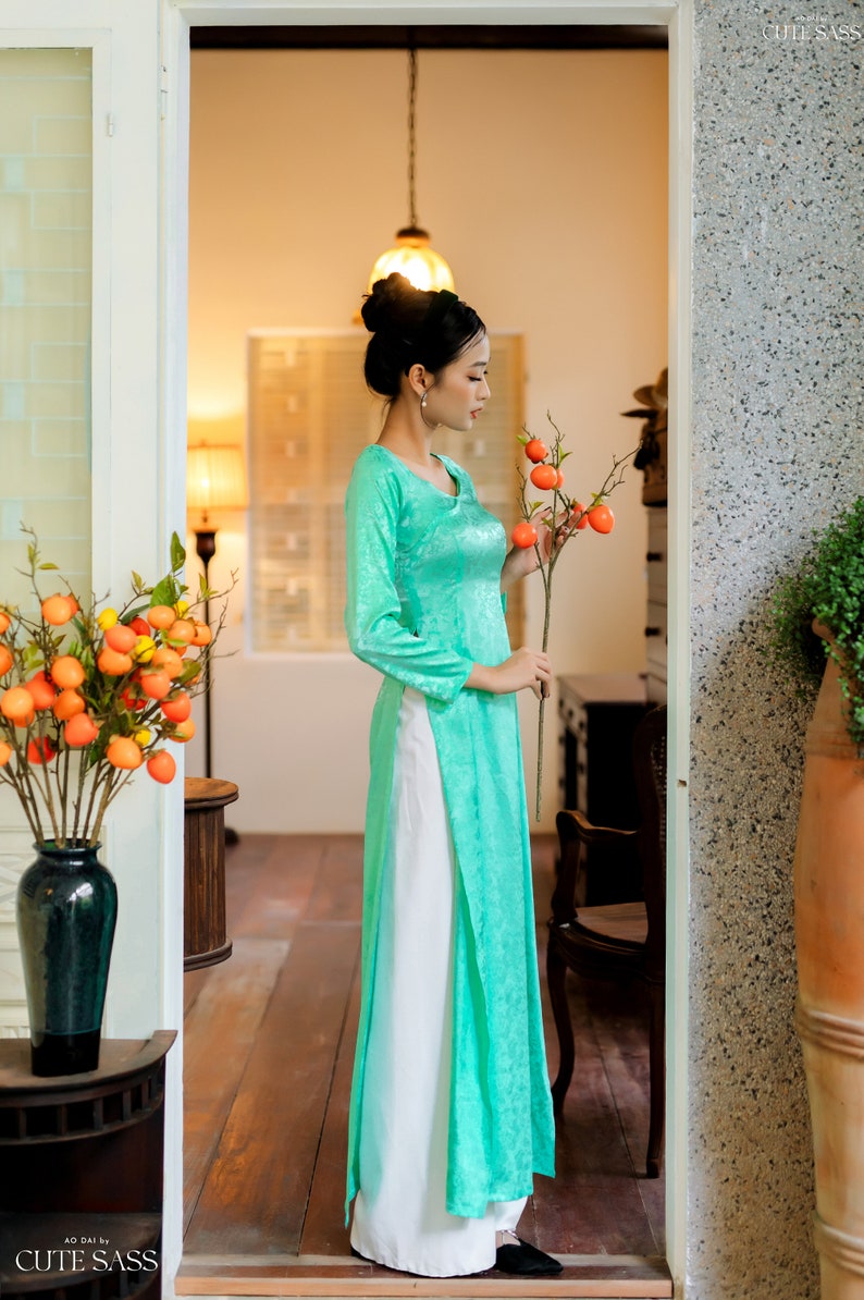Op de afbeelding: Een vrouw in een turquoise zijden jurk met een witte rok en een tak met oranje fruit vasthoudt. De jurk heeft een traditionele Vietnamese stijl.