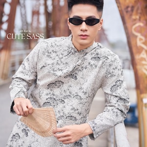 May include: A man wearing a white long-sleeved shirt with a black floral pattern and black sunglasses. He is holding a light brown fan in his right hand. The shirt has the text "AO DAI by CUTE SASS" on the front.