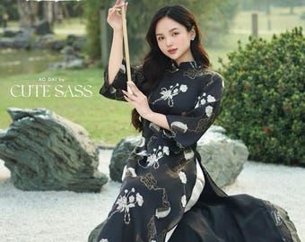 Pode incluir: Uma mulher vestindo um vestido preto com padrão floral branco, gola alta e mangas compridas. Ela está segurando um guarda-chuva branco e sentada em uma grande rocha. O texto "AO DAI by CUTE SASS" é visível na imagem.