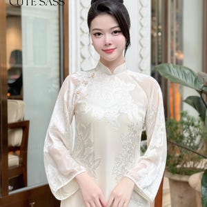 May include: A white traditional Vietnamese dress with floral embroidery, called an Ao Dai, with a high collar and long sleeves. The dress is worn by a woman with her hair pulled back in a bun. The text "AO DAI by CUTE SASS" is visible at the top of the image.