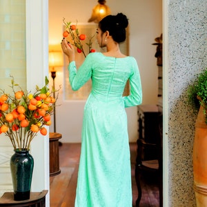 Op de afbeelding: Een vrouw in een lichtgroene, lange jurk met lange mouwen en een witte onderrok staat in een deuropening. Ze houdt een tak vast met kleine oranje vruchten.