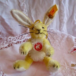 May include: A yellow plush bunny rabbit with long ears, black whiskers, and a stitched mouth. The bunny has a red and white tag that says "Made in Germany". A yellow tag is attached to the ear.