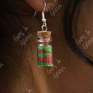 May include: A pair of earrings featuring small glass bottles filled with red and green glitter. The bottles have cork stoppers and are attached to silver earring hooks.
