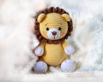 Puede incluir: Un juguete de peluche de león tejido a crochet con una melena marrón, un cuerpo amarillo y patas blancas. El león tiene una cara linda con ojos negros y una nariz rosa.