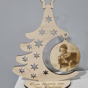 Può includere: Un ornamento per albero di Natale in legno con un'incisione a forma di mezzaluna e stelle. Una foto rotonda di un bambino è attaccata all'ornamento. La base dell'ornamento ha il testo "Buon Natale dai tuoi Padrini".