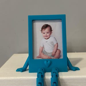 Könnte beinhalten: Türkisfarbener Bilderrahmen mit einem Babyfoto. Der Rahmen hat Arme, Füße und Reißverschlussdetails. Unten befindet sich ein Herz. Das Baby trägt ein weißes Oberteil und gestreifte Shorts.