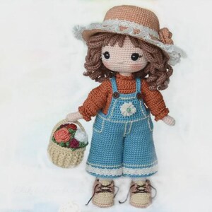 May include: A handmade crochet doll wearing a brown hat with a white lace trim, a rust-colored sweater, and blue overalls. The doll holds a small basket filled with colorful flowers and has brown curly hair. The doll is approximately 10 inches tall.