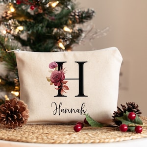 May include: A white canvas makeup bag with a floral design and the letter "H" in black. The name "Hannah" is printed below the letter.