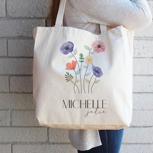 May include: Cream-colored tote bag featuring a floral design with purple, pink, orange, and yellow flowers. The bag has the name "MICHELLE jolie" printed in black. The bag is carried by a person.