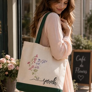 A woman in a pink dress carrying a white tote bag with a floral design and the name "Sarah" printed on it, standing in front of a cafe sign.