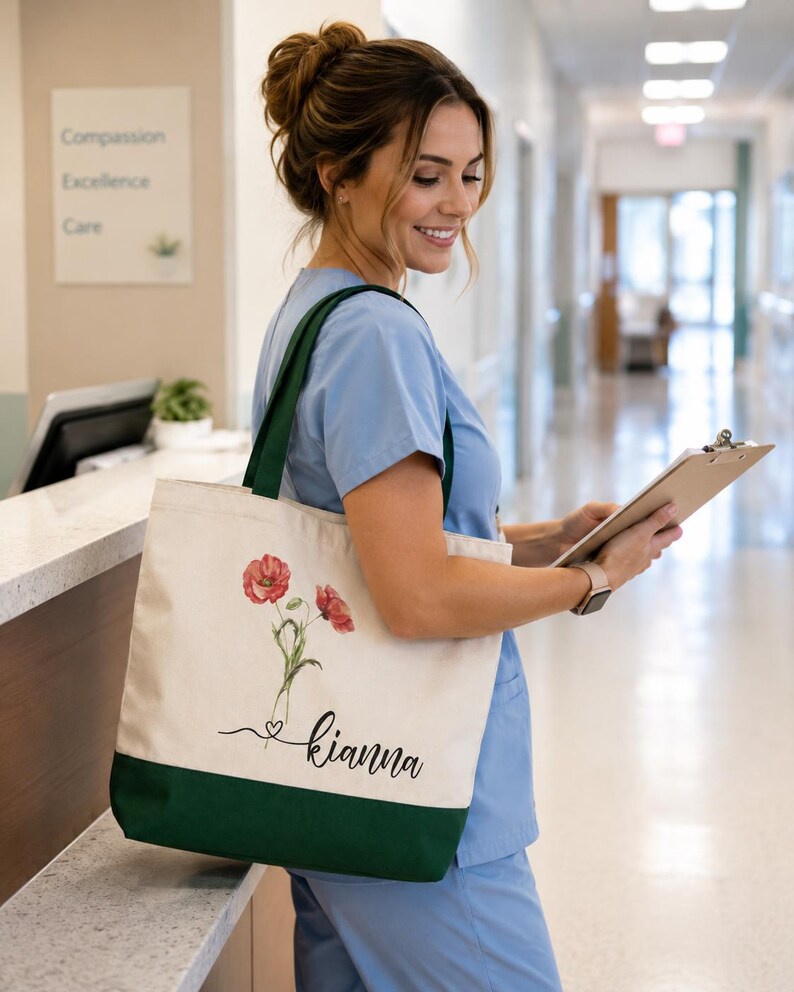 A smiling woman in blue scrubs stands in a hallway, holding a clipboard and wearing a tote bag with a floral design.