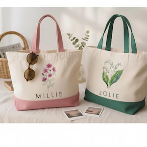 May include: Two canvas tote bags with floral designs. One bag is pink and features the name "Millie" and a pink flower graphic. The other bag is green and features the name "Jolie" and a lily of the valley graphic.