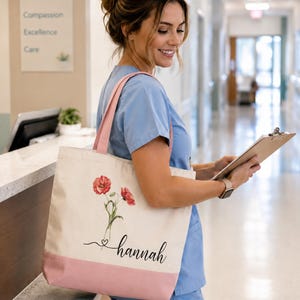 A smiling woman in blue scrubs is standing in a hallway, holding a clipboard and a bag with a floral design.