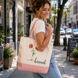 A woman is standing on a sidewalk, smiling and carrying a pink and white tote bag with a floral design.