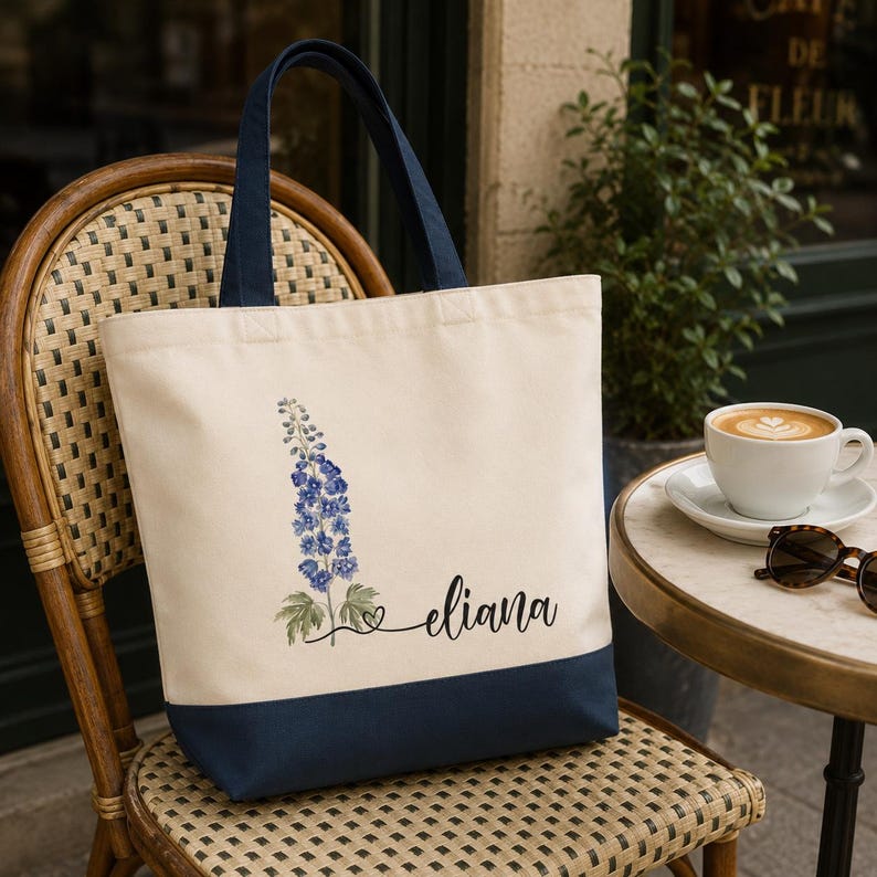 A white tote bag with a blue trim and a floral design, sitting on a woven chair next to a small table with a cup of coffee and sunglasses.