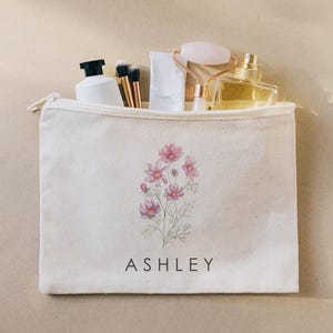 May include: A cream-colored canvas zipper pouch featuring a printed illustration of pink cosmos flowers and the name ASHLEY in black capital letters. The pouch is filled with a white bottle, two makeup brushes, a white tube, a facial roller, and a perfume bottle.