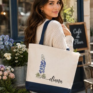 A woman with long brown hair is standing in front of a storefront, holding a white tote bag with a blue trim and a flower design.