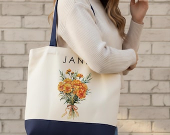 May include: Cream-colored tote bag with a navy blue base and straps. The bag features the word "JAN." above a watercolor illustration of orange flowers tied with a ribbon. The bag is held by a person.