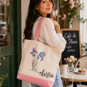A young woman with long brown hair is standing in front of a cafe, holding a white tote bag with a pink trim and a floral design.