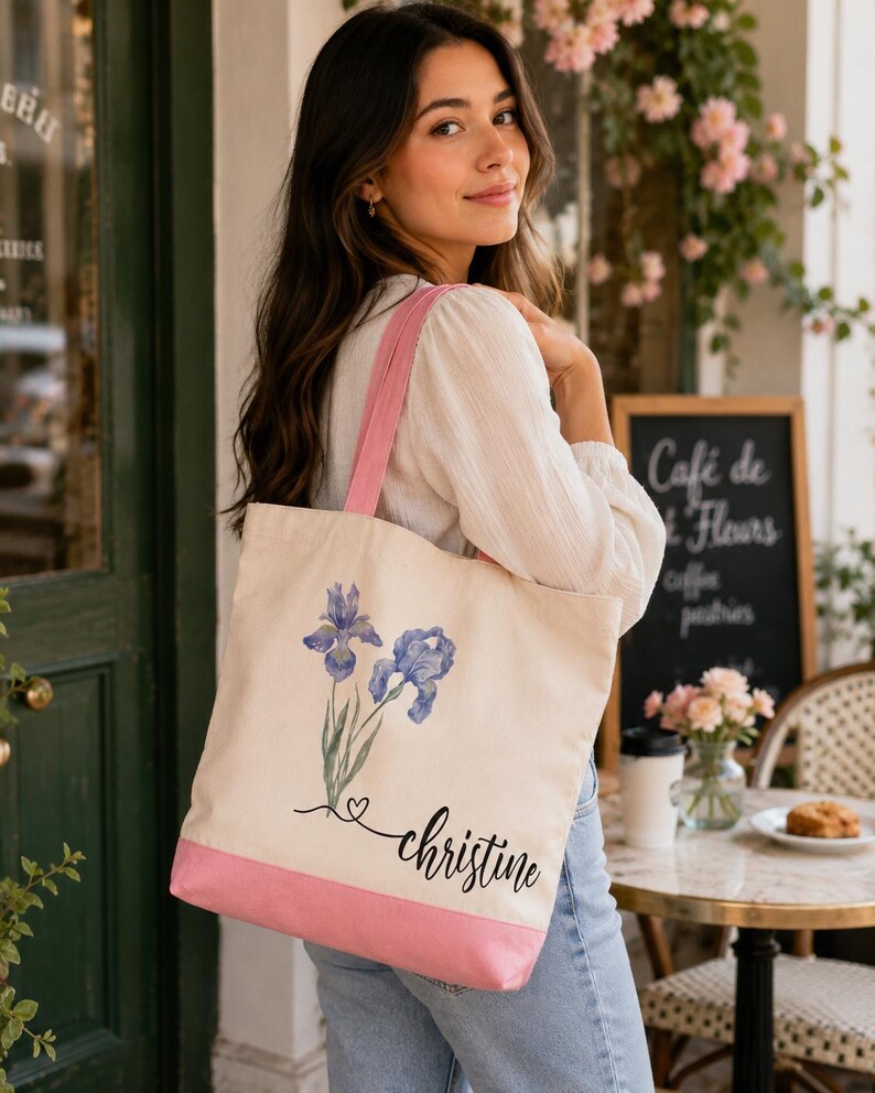 A young woman with long brown hair is standing in front of a cafe, holding a white tote bag with a pink trim and a floral design.