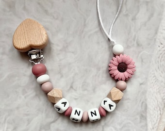 Könnte beinhalten: Ein Holz-Schnuller in Herzform mit einer rosa und weißen Silikonperlenkette. Die Kette hat Holzperlen mit den Buchstaben "ANNA" und eine rosa Silikonblume mit braunem Zentrum.