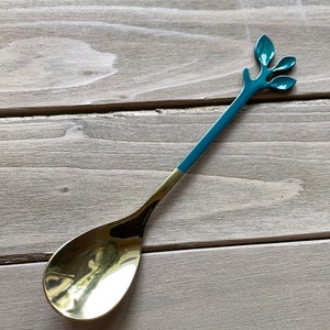 May include: A gold and teal spoon with a leaf design on the handle. The spoon is on a wooden surface.