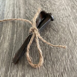 May include: A bundle of three dark metal nails tied together with natural twine. The nails have a rectangular shape with a rounded head. The twine is knotted in a decorative bow.
